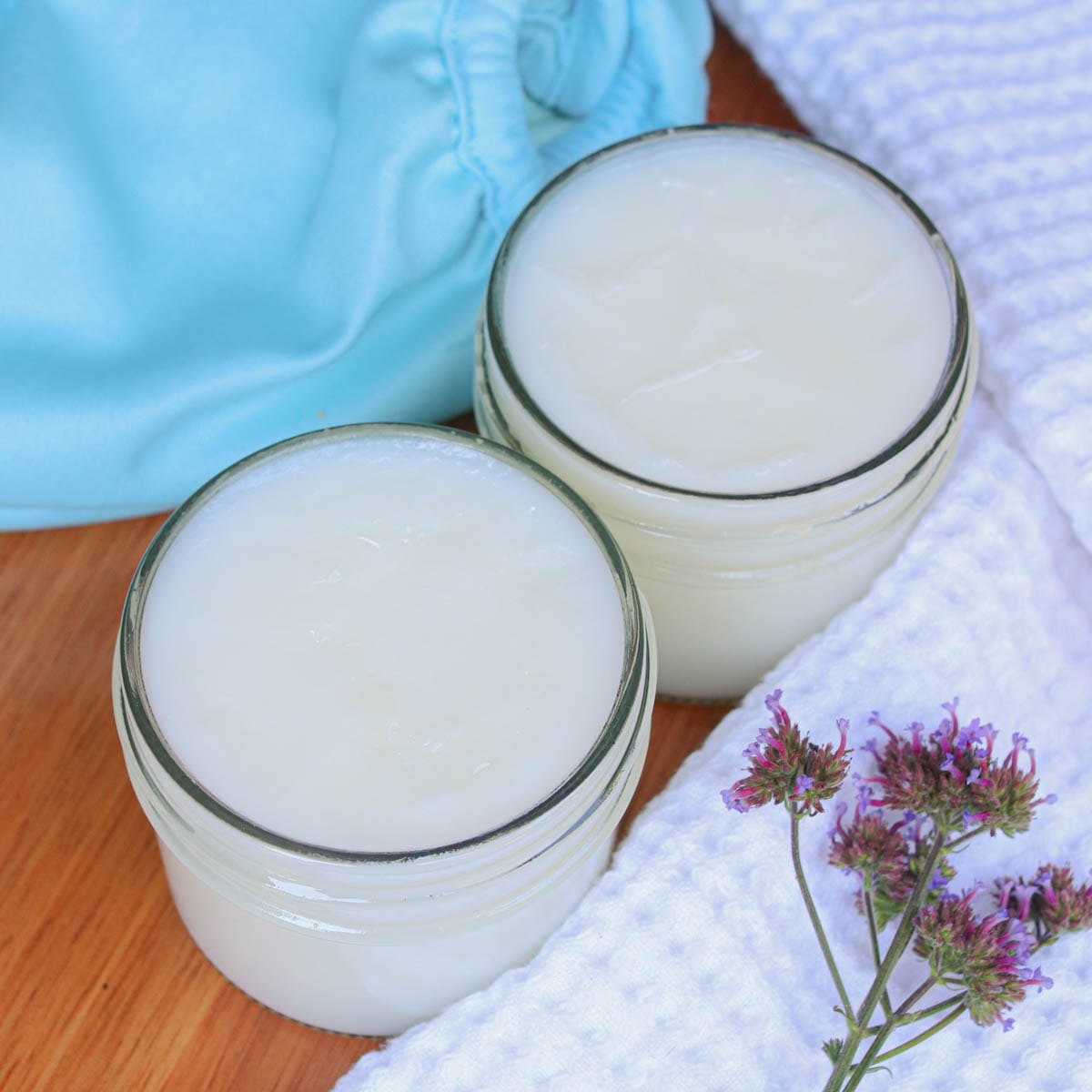 Two glass jars of baby balm on a wooden surface with a purple flower and a white cloth.