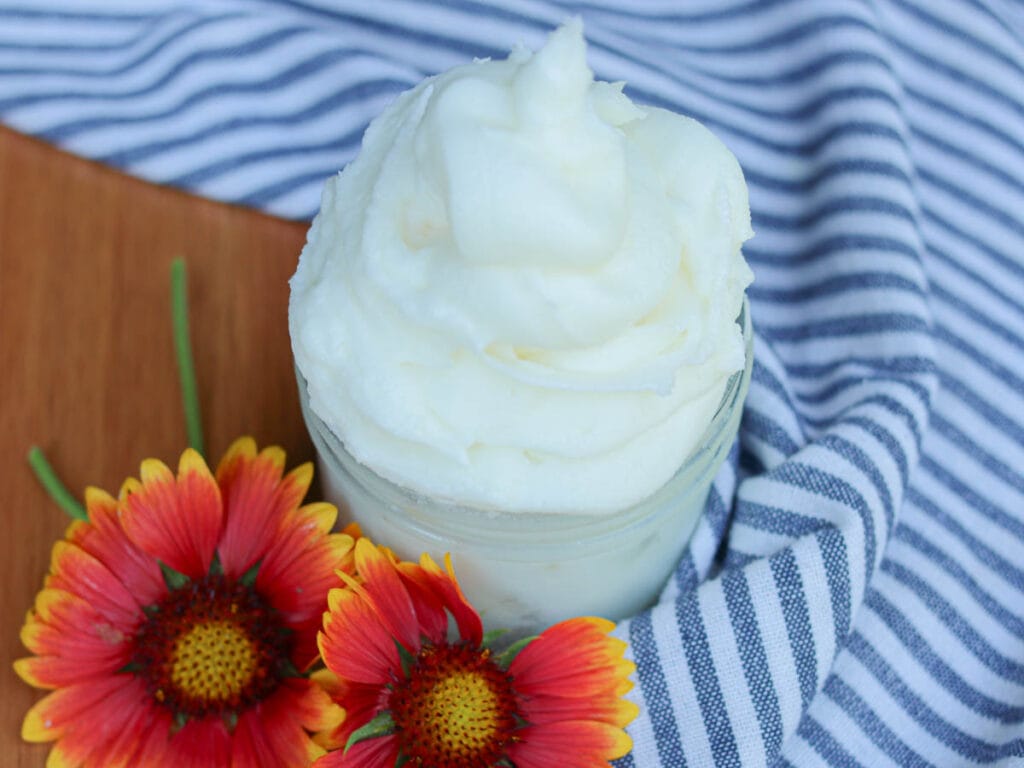 Fluffy white whipped body butter in a glass jar on a wooden surface with a striped cloth and flowers.