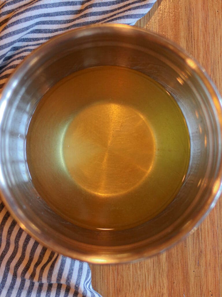 Melted body butter ingredients in a stainless steel bowl on a wooden surface with a striped cloth.