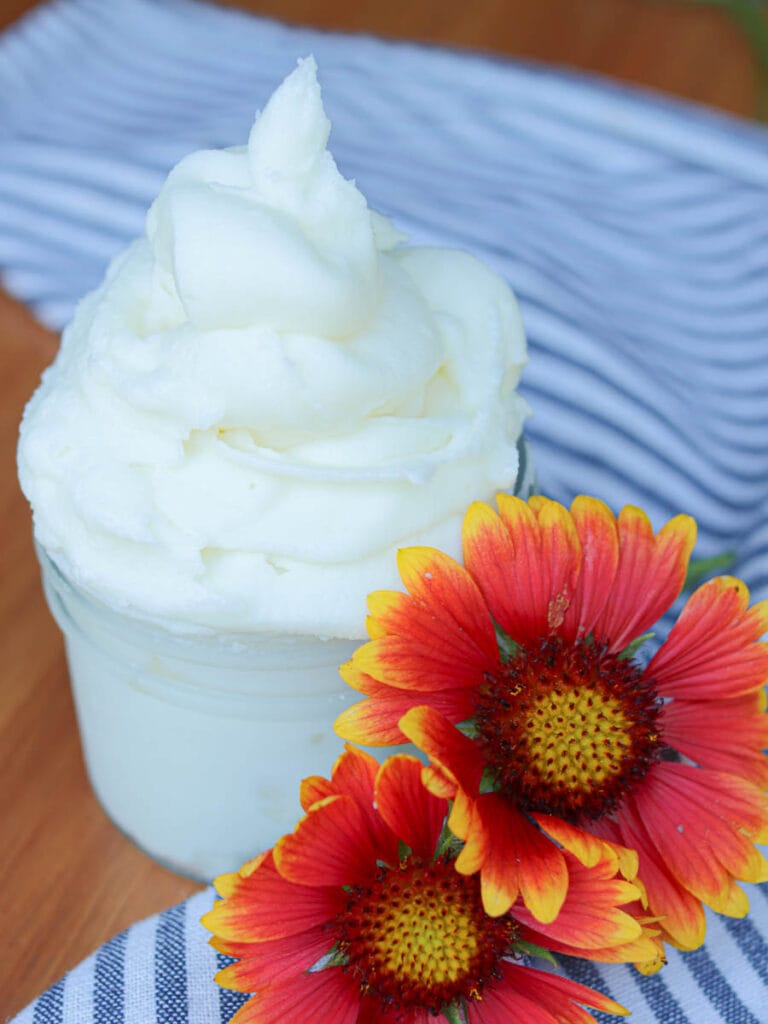 Fluffy whipped body butter in a glass jar on a wooden surface with a striped cloth and flowers.