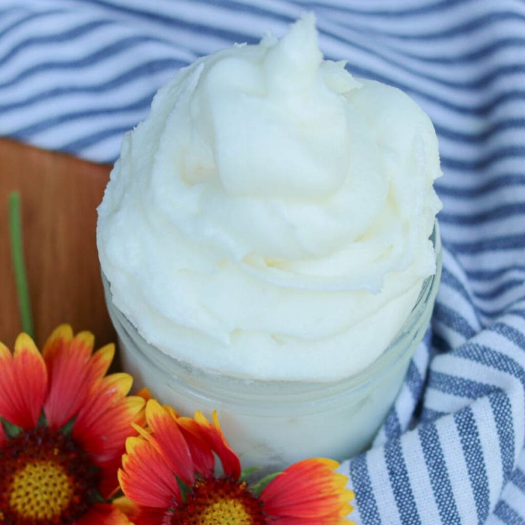 Fluffy whipped body butter in a glass jar on a wooden surface with a striped cloth and fresh flowers.