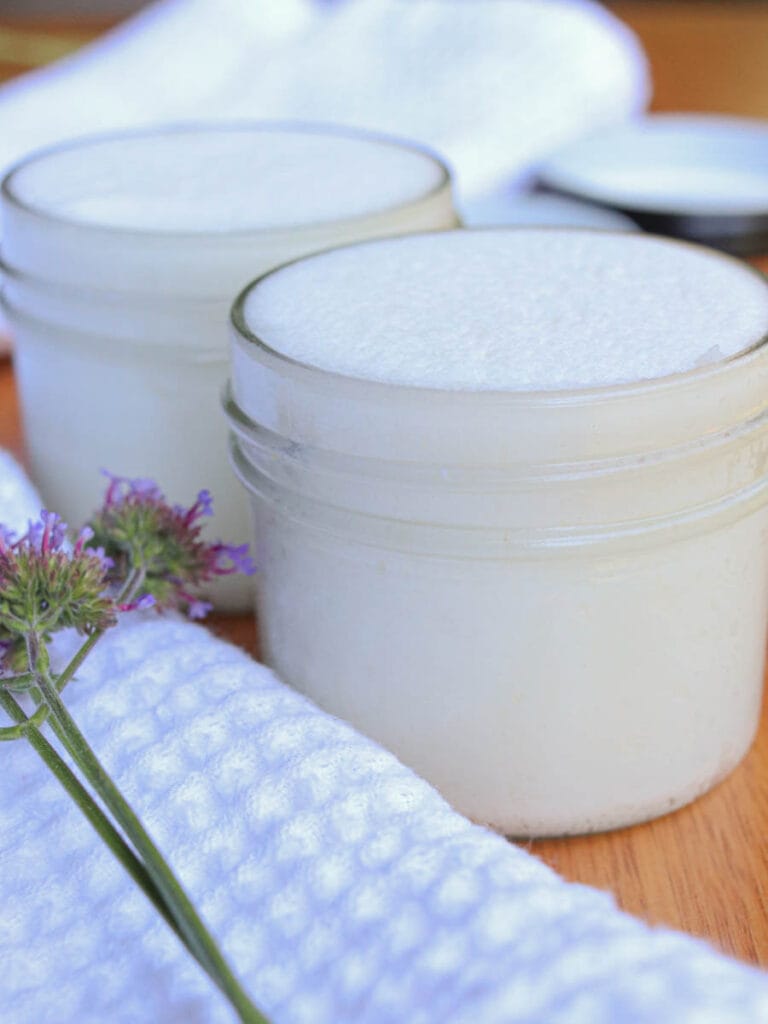 Two glass jars filled with DIY sugar scrub on a wooden surface next to a white cloth.