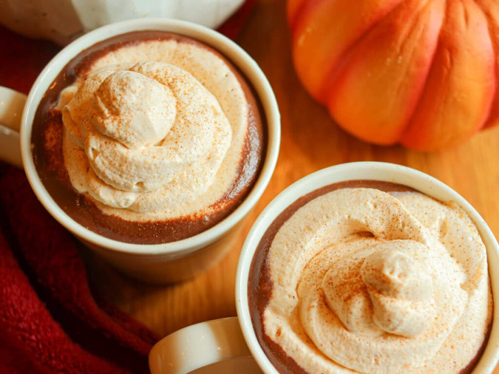 Two white mugs full of hot chocolate with pumpkin spice topped with pumpkin spice whipped cream on a wooden table with pumpkins.