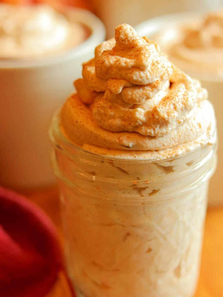Glass jar filled with pumpkin spice whipped cream with mugs of hot chocolate in the background.