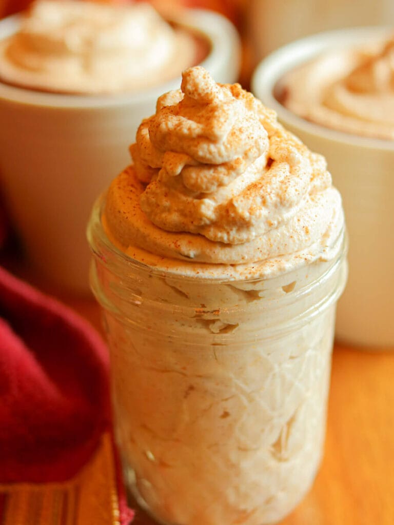 Glass jar filled with fluffy pumpkin spice whipped cream with cups of hot chocolate in the background.