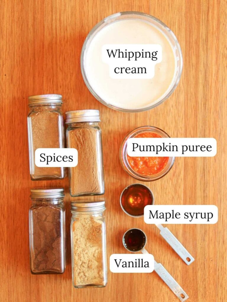 Labeled ingredients on a wooden table including whipping cream, spices, maple syrup, vanilla, and pumpkin puree.