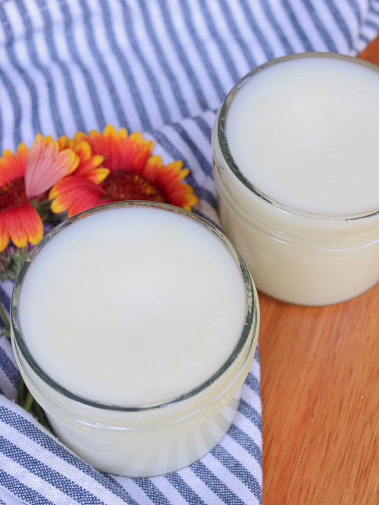 Two open glass jars of tallow balm on a wooden surface with a striped cloth and orange flowers.