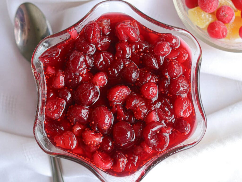 Top view of cranberry compote in a glass dish next to a spoon on a white tablecloth.