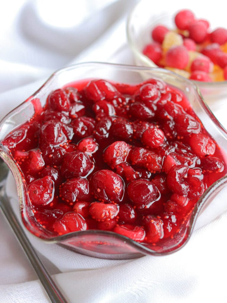 Closeup of cranberry compote in a glass dish next to a spoon and bowl of cranberries and crystalized ginger on a white tablecloth.