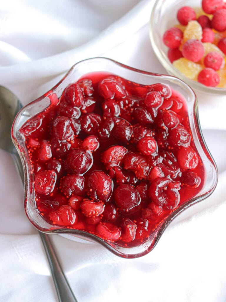 Cranberry compote in a glass dish next to a spoon and bowl of cranberries and crystalized ginger on a white tablecloth.