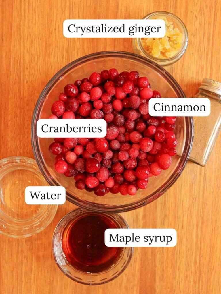 Labeled ingredients on a wooden table including cranberries, maple syrup, crystalized ginger, cinnamon, and water.