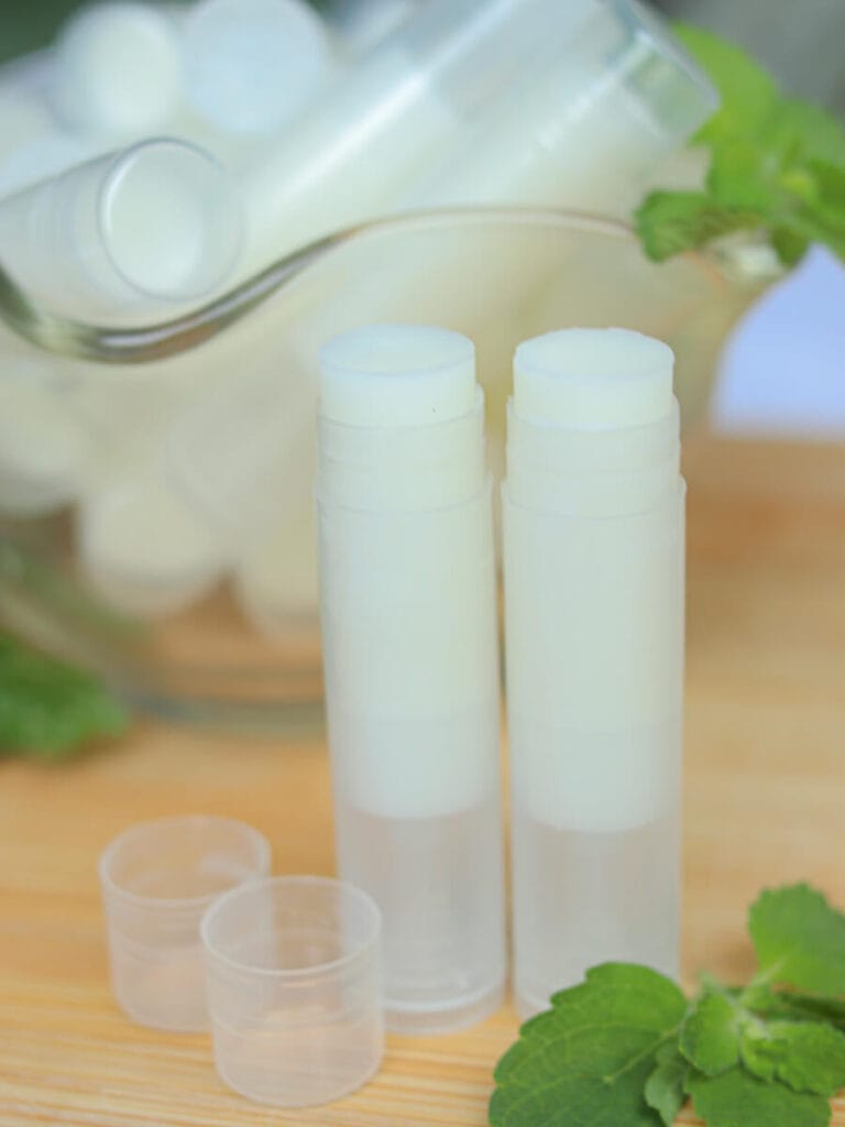 Two open tubes of homemade lip balm sitting on a wooden surface with more lip balm in a bowl in the background.