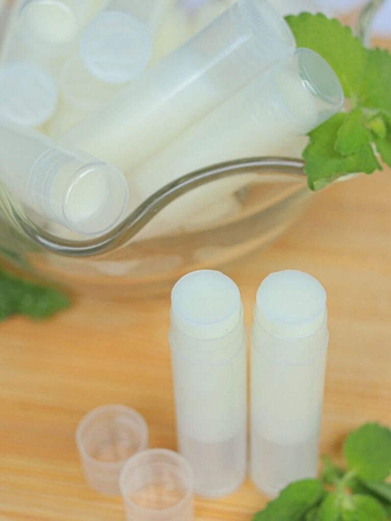 Two open tubes of homemade lip balm sitting on a wooden surface with more lip balm in the background.