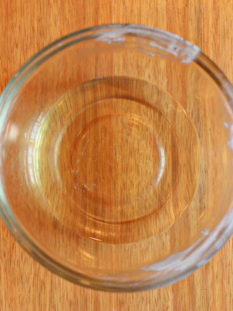Melted tallow, melted beeswax, sweet almond oil, and essential oil in a glass mixing bowl on a wooden table.