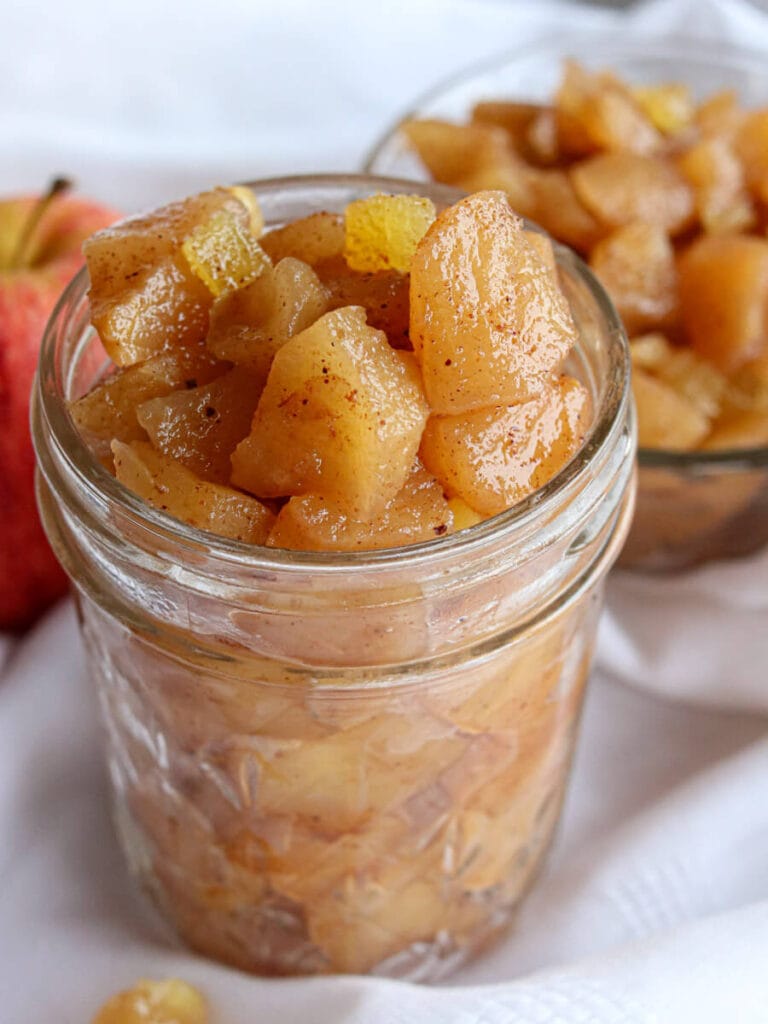 Closeup of apple compote heaped in a glass jar on a white surface.