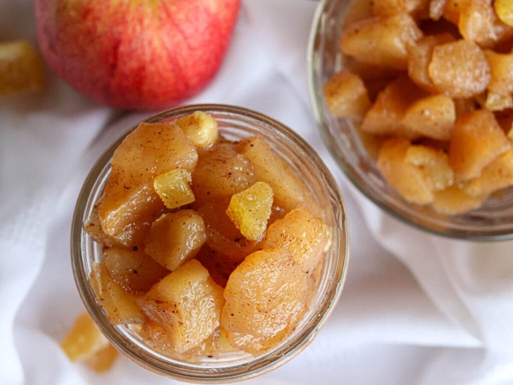 Top view of a glass jar of apple compote beside an apple and a bowl with more compote.