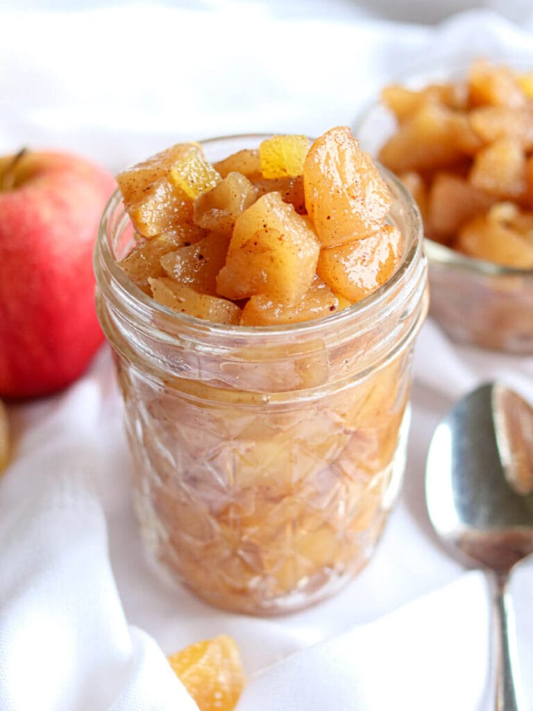 Apple compote in a glass jar on a white tablecloth beside a spoon with an apple in the background.