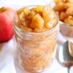 Closeup of apple compote in a glass jar on a white tablecloth.