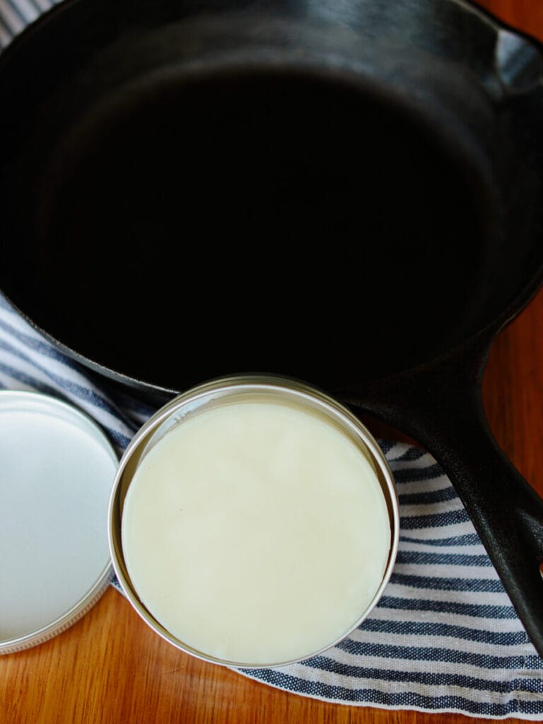 A metal tin with a bar of cast iron seasoning wax beside a cast iron skillet.