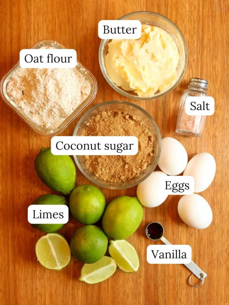Labeled ingredients on a wooden table including oat flour, butter, coconut sugar, salt, limes, eggs, and vanilla.