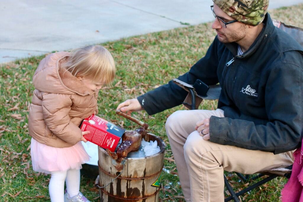 Toddler pouring rock salt into an ice cream maker that a man is churning.