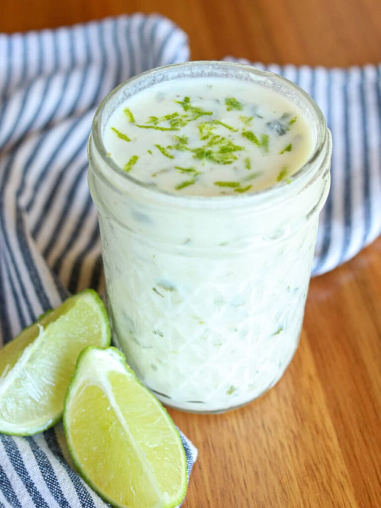 Lime crema topped with lime zest in a glass mason jar on a wooden table beside two lime slices.