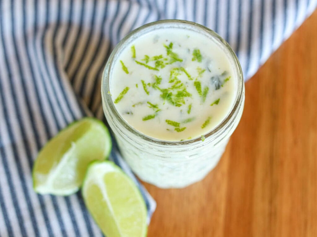 Top view of lime crema garnished with lime zest in a glass jar on a wooden table beside two lime slices.