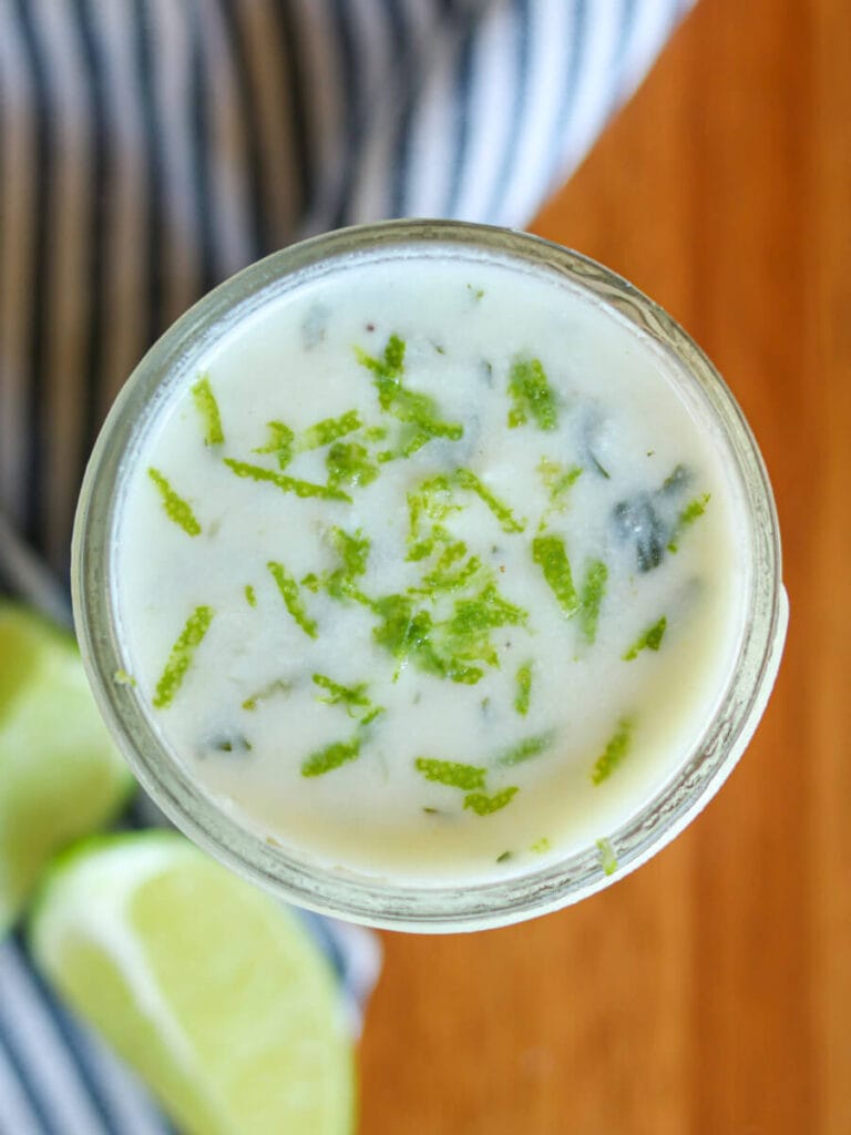 Lime crema in a glass jar on a wooden table beside lime slices.