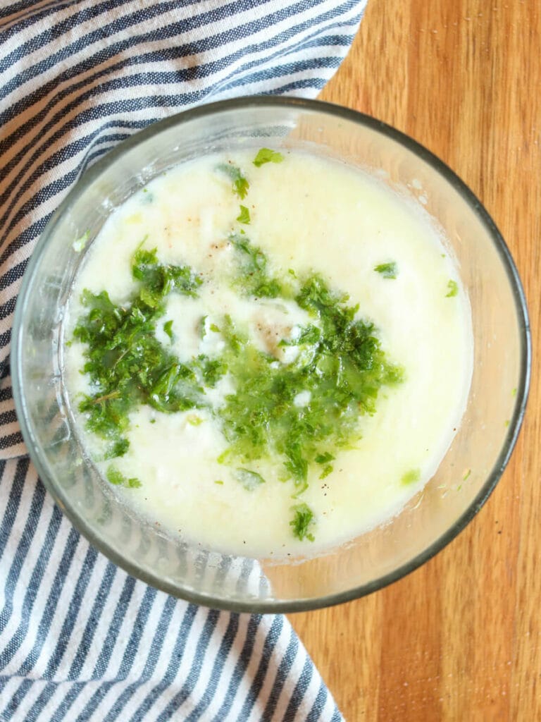 Yogurt, lime juice, and other ingredients in a mixing bowl on a wooden table beside a striped cloth napkin.