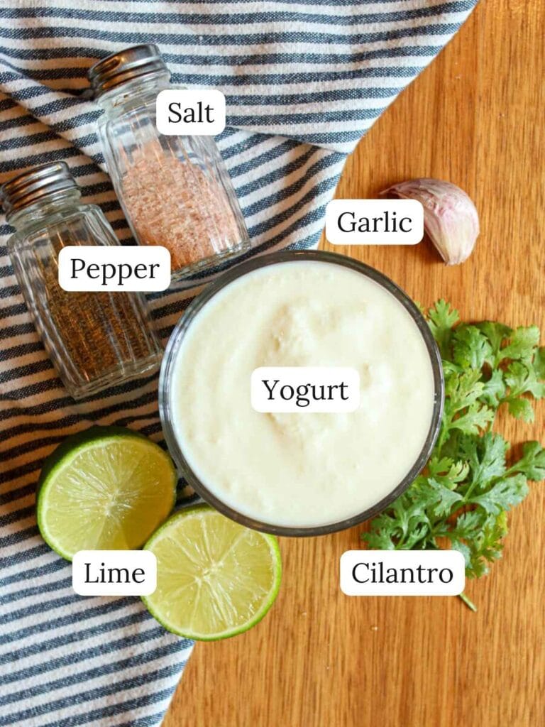Labeled ingredients on a wooden surface including yogurt, garlic, salt, pepper, cilantro, and lime.