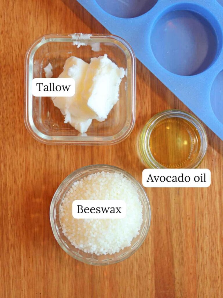 Labeled ingredients on a wooden surface including tallow, beeswax, and avocado oil.