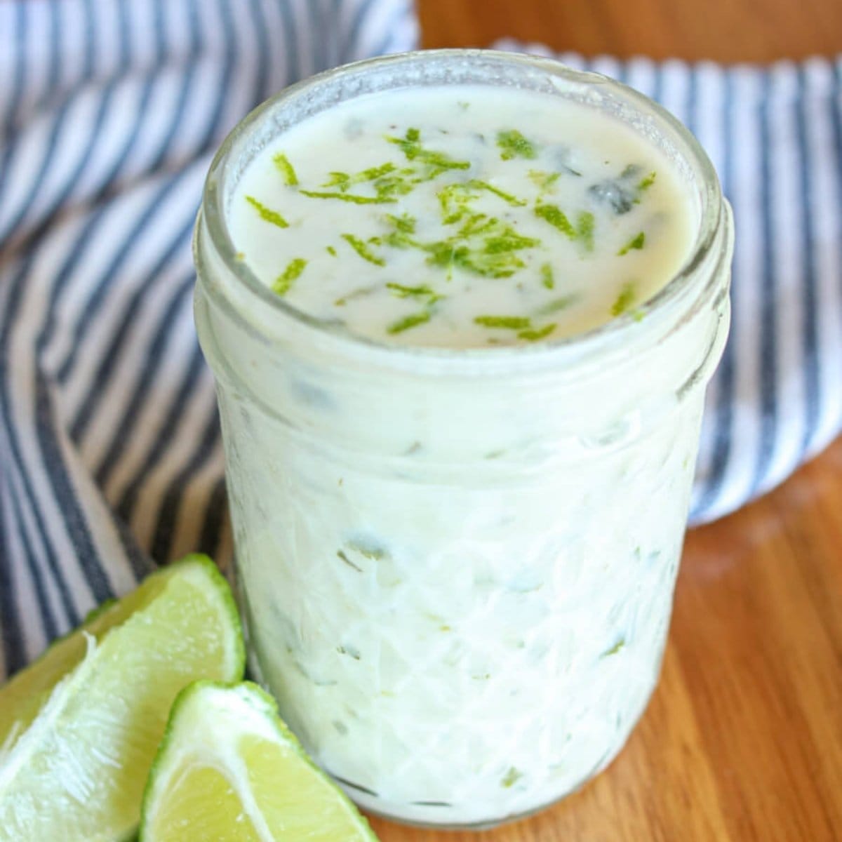 Closeup of lime crema topped with lime zest in a glass mason jar on a wooden table with fresh lime slices.