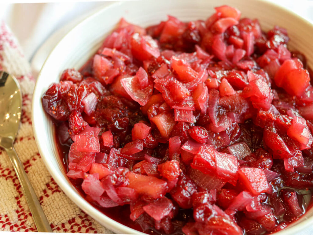 A serving bowl with apple cranberry chutney beside a serving spoon and kitchen towel.