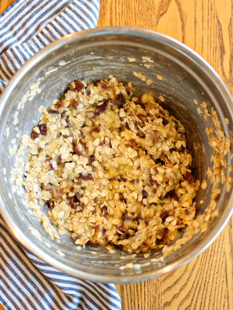 Mixing oats, chopped dates, bananas, and other ingredients in a bowl on a wooden surface.