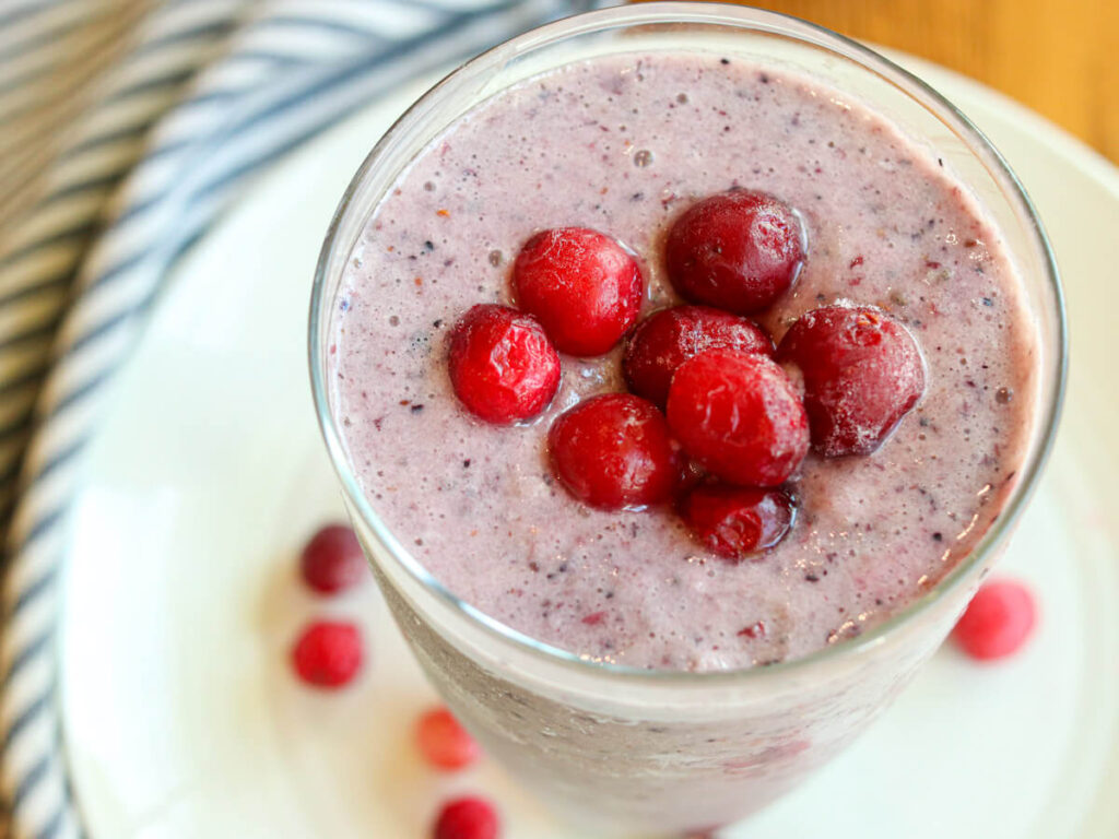Top view of a cranberry smoothie in a glass on a white plate topped with frozen cranberries.