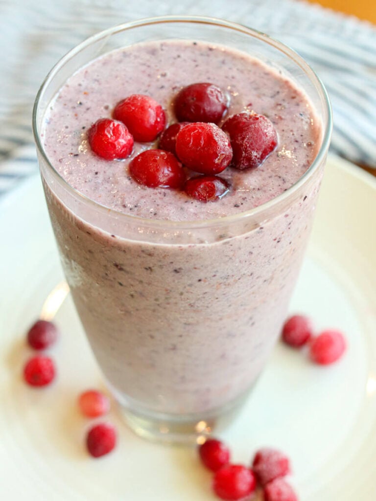 Bright blueberry cranberry smoothie in a tall glass topped with frozen cranberries on a white plate.