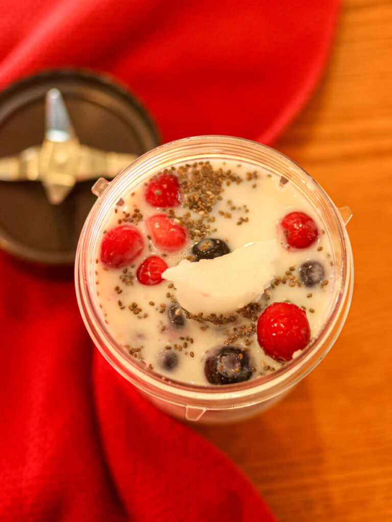 Smoothie cup with milk, cranberries, coconut oil, and other smoothie ingredients beside a red kitchen towel.