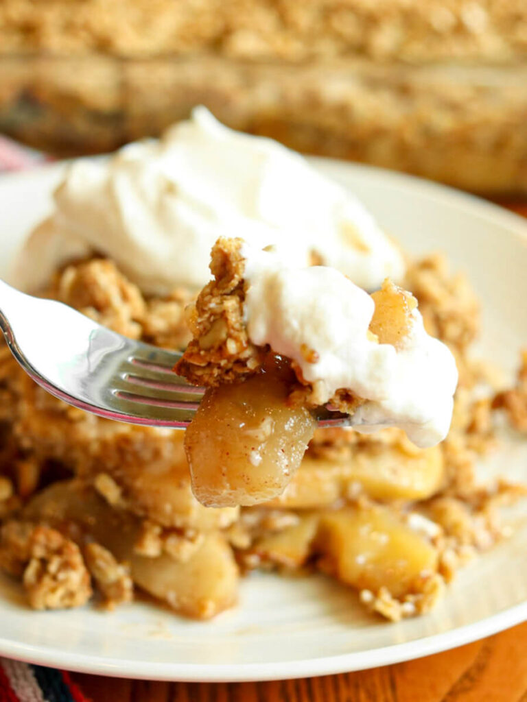 Closeup of a bite of apple crisp with whipped cream on a fork with a large piece on a plate in the background.