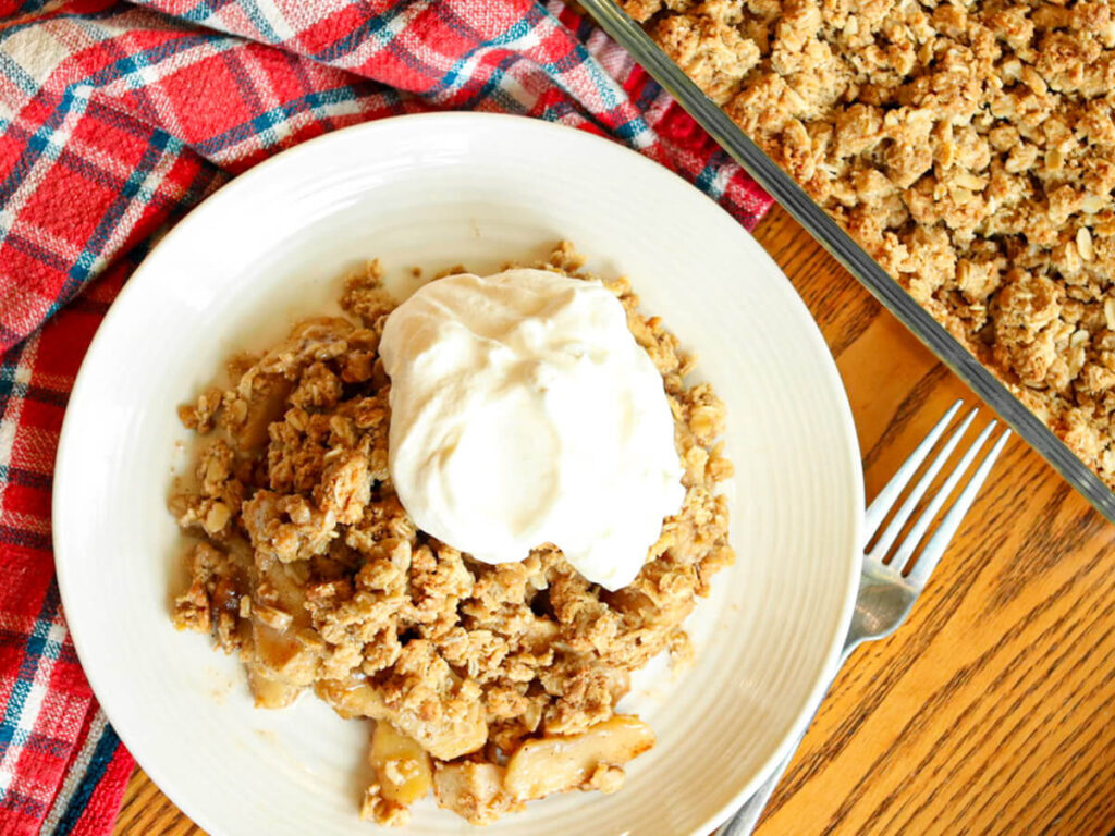 Top view of apple crisp and whipped cream on a plate and in a baking dish surrounded by a kitchen towel on a wood table.