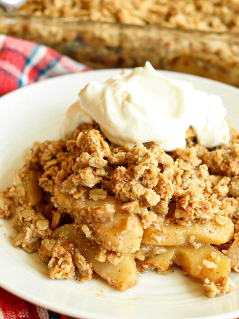 A heaping serving of warm apple crisp topped with whipped cream on a plate with pan of apple crisp in the background.