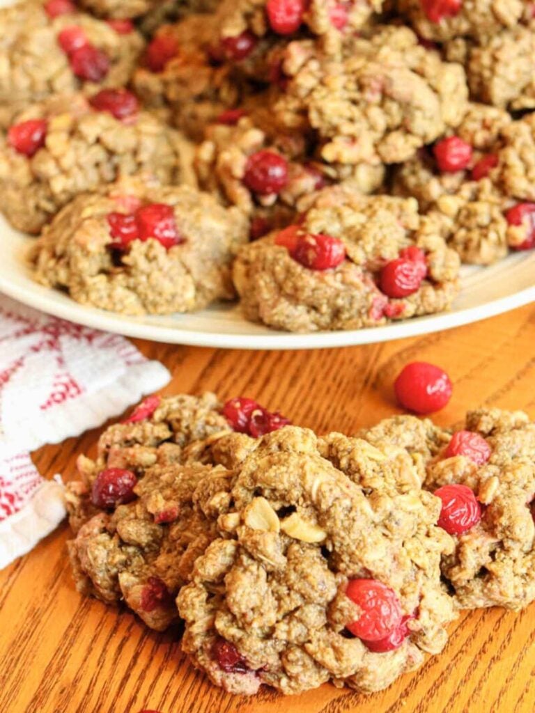 Three cookies on a wooden table with a few frozen cranberries around them and a plate of more cookies in the background.
