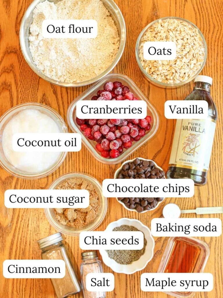 Labeled ingredients on a wood background including oat flour, oats, coconut oil, cranberries, vanilla, coconut sugar, chocolate chips, cinnamon, chia seeds, salt, baking soda, and maple syrup.