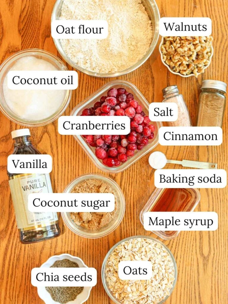 Labeled ingredients on a wood table including oat flour, oats, coconut oil, cranberries, vanilla, coconut sugar, walnuts, cinnamon, chia seeds, salt, baking soda, and maple syrup.