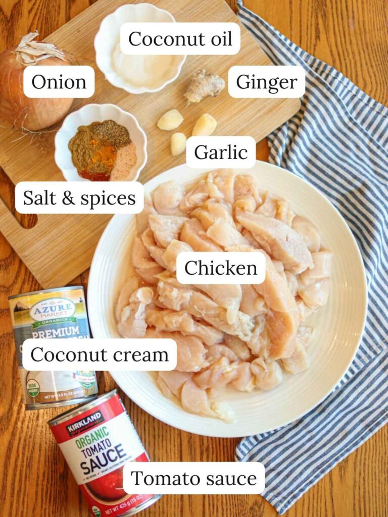 Labeled ingredients on a wooden table with a cloth napkin including chicken, tomato sauce, coconut cream, salt and spices, coconut oil, ginger, onion, and garlic.