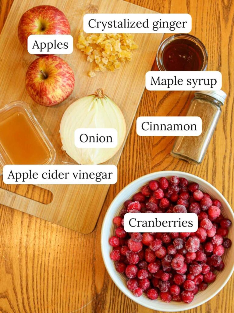 Labeled ingredients on a wooden table and cutting board including apples, onion, crystalized ginger, apple cider vinegar, cinnamon, maple syrup, and cranberries.