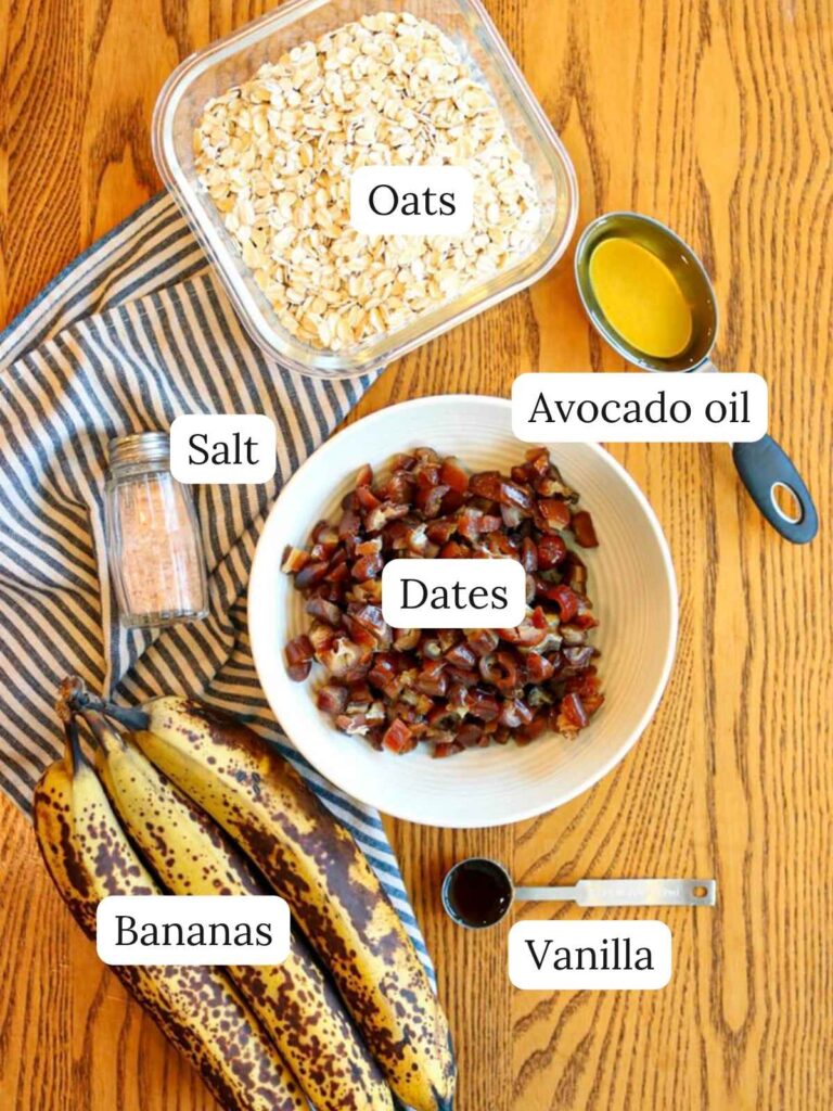 Labeled ingredients on a wooden table including oats, salt, avocado oil, dates, bananas, and vanilla.