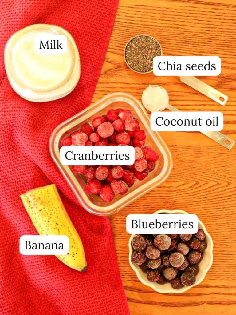 Labeled ingredients on a wooden table including milk, cranberries, chia seeds, coconut oil, banana, and blueberries.