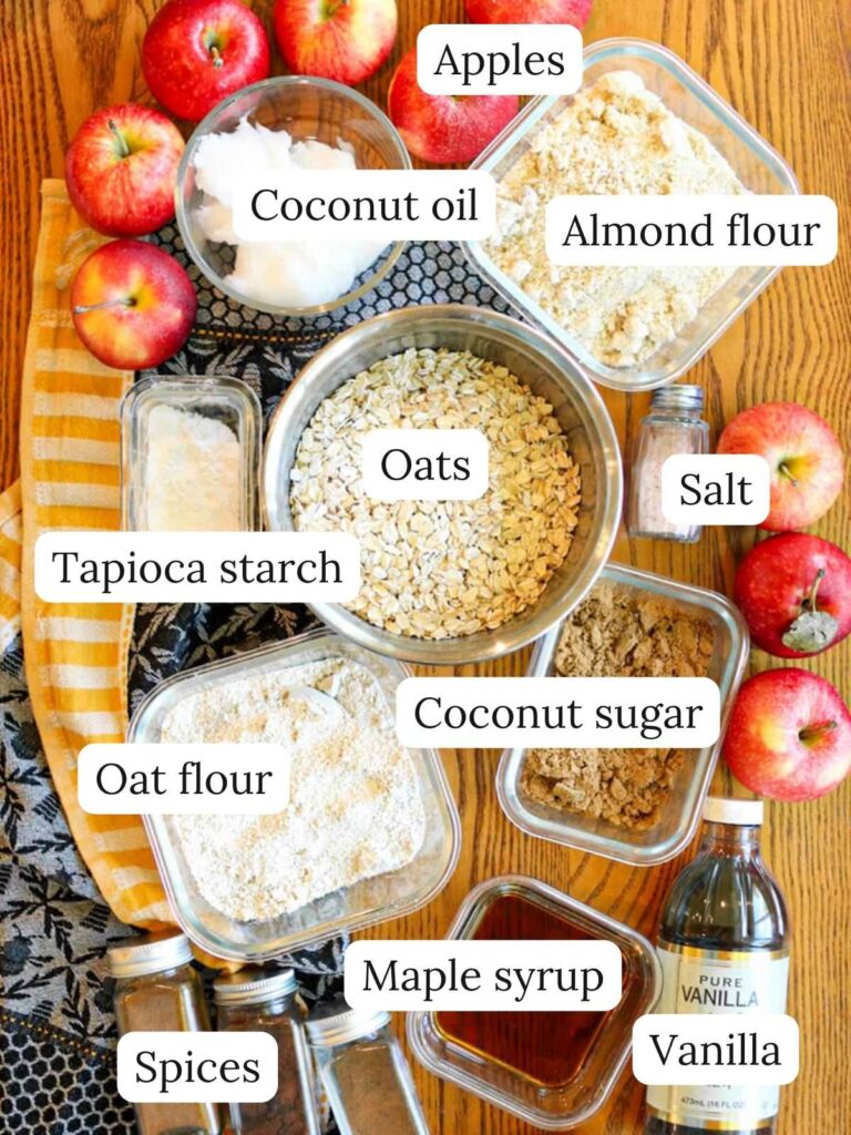 Labeled ingredients on a wooden table with a navy and yellow dish towel including apples, coconut oil, almond flour, oats, tapioca starch, salt, oat flour, coconut sugar, spices, maple syrup, and vanilla.