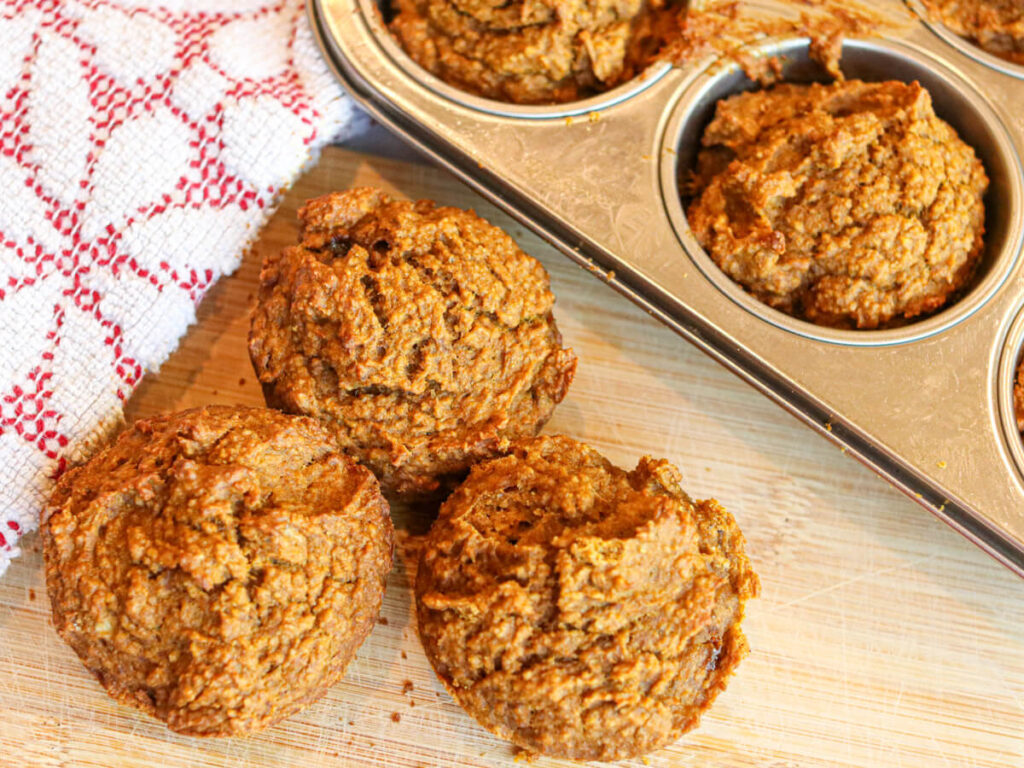 Three healthy banana pumpkin muffins on a wooden table beside a pan with more muffins.