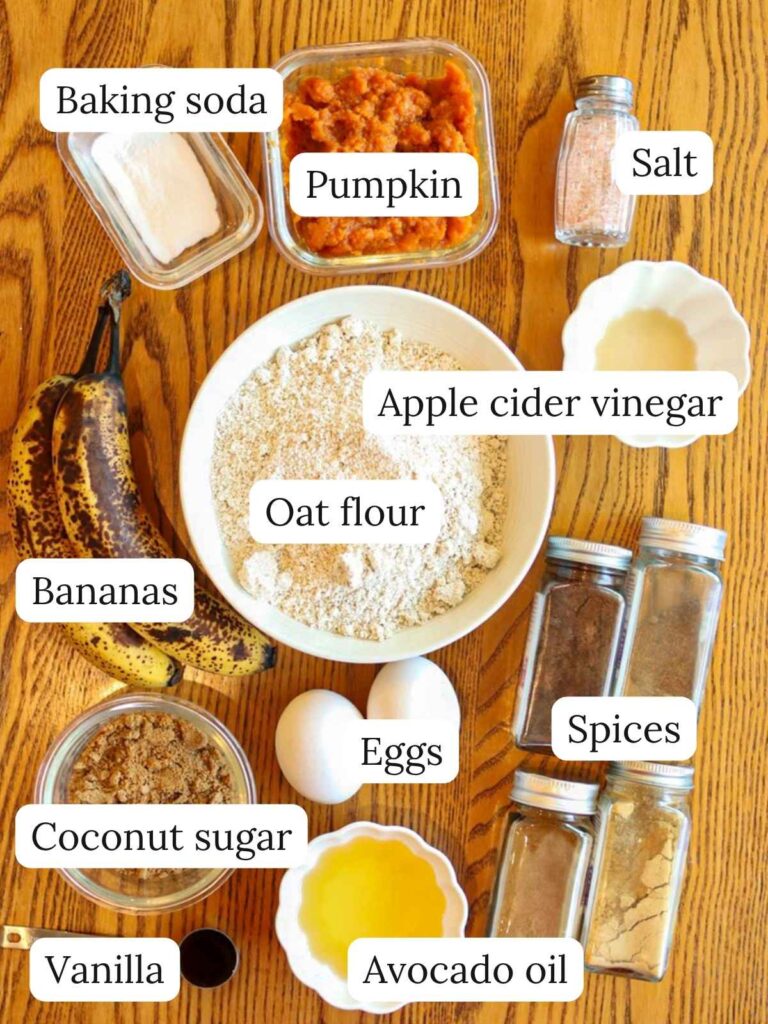 Labeled muffin ingredients on a wooden table including baking soda, pumpkin, salt, apple cider vinegar, oat flour, bananas, eggs, coconut sugar, avocado oil, spices, and vanilla.